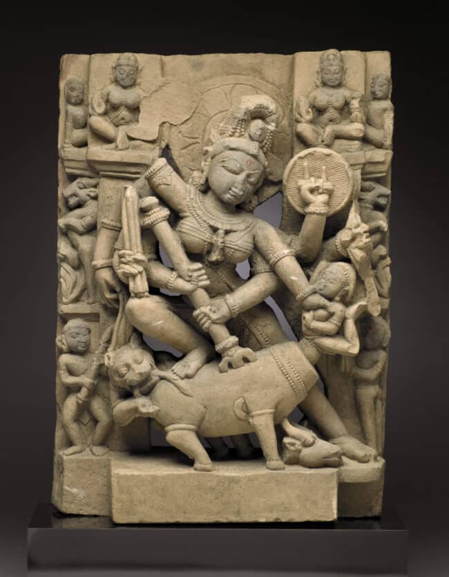 Ancient stone sculpture depicting a goddess holding a shield and weapon, standing over a buffalo, surrounded by smaller figures.