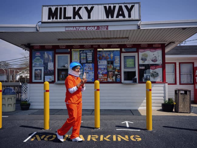 Person in orange jumpsuit and astronaut helmet looking at an ice cream cone in front of a storefront with a sign that says "MILKY WAY" at the rooftop