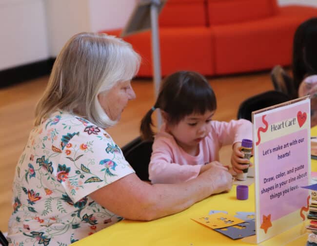 A small child is being helped with her artwork by an adult at the Art Cart table