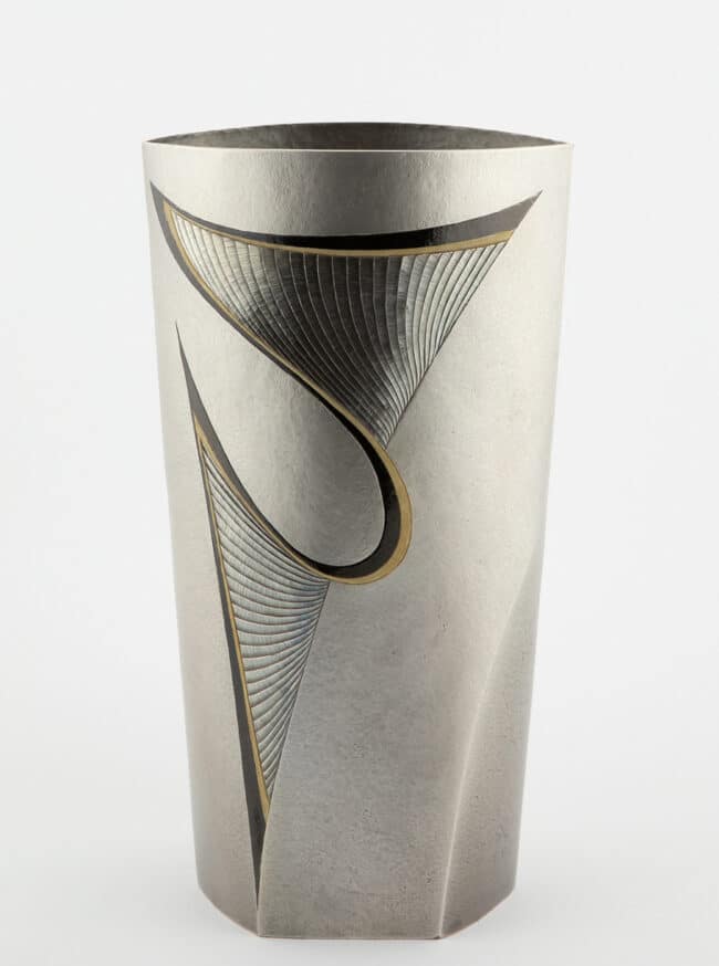 A front-facing shot of a cylindrical silver vase with curvilinear gold inlay patterning