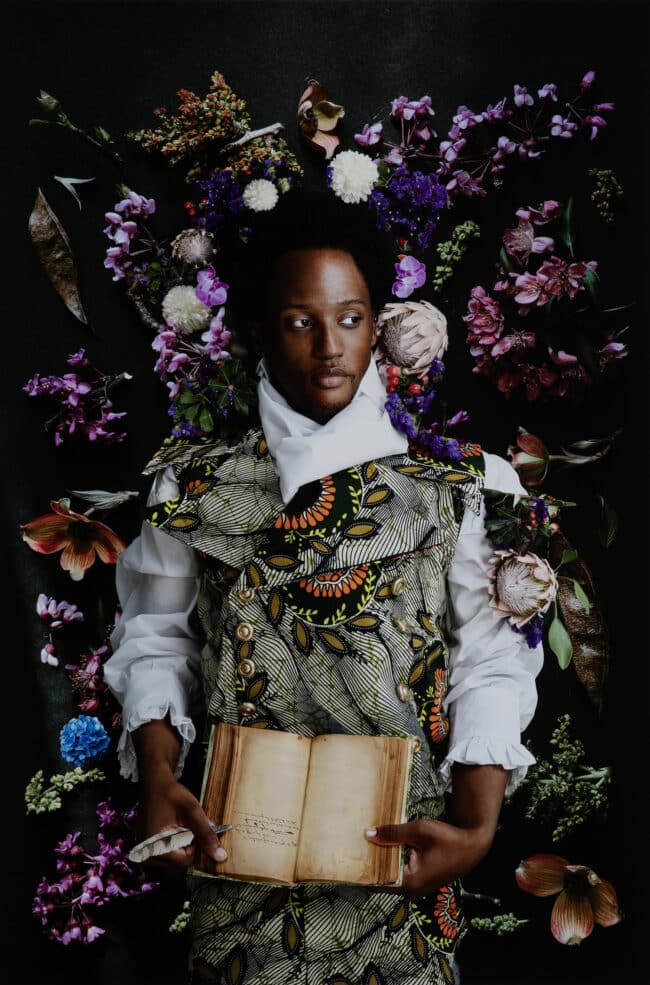 Photograph portrait of a Black man looking off to the right of the frame, surrounded by flowers, holding a quill pen and journal