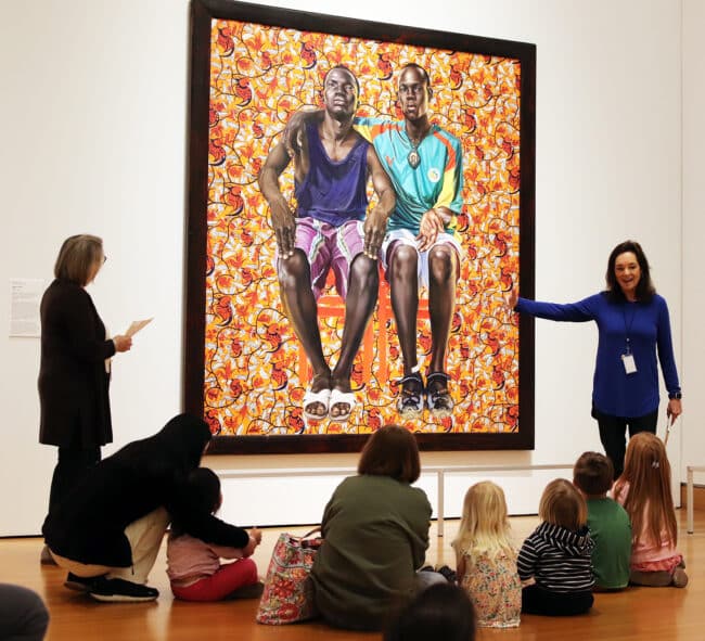 young visitors and their caregivers sitting on the floor in the contemporary gallery listening to a presentation by docents in front of a Kehinde Wiley painting