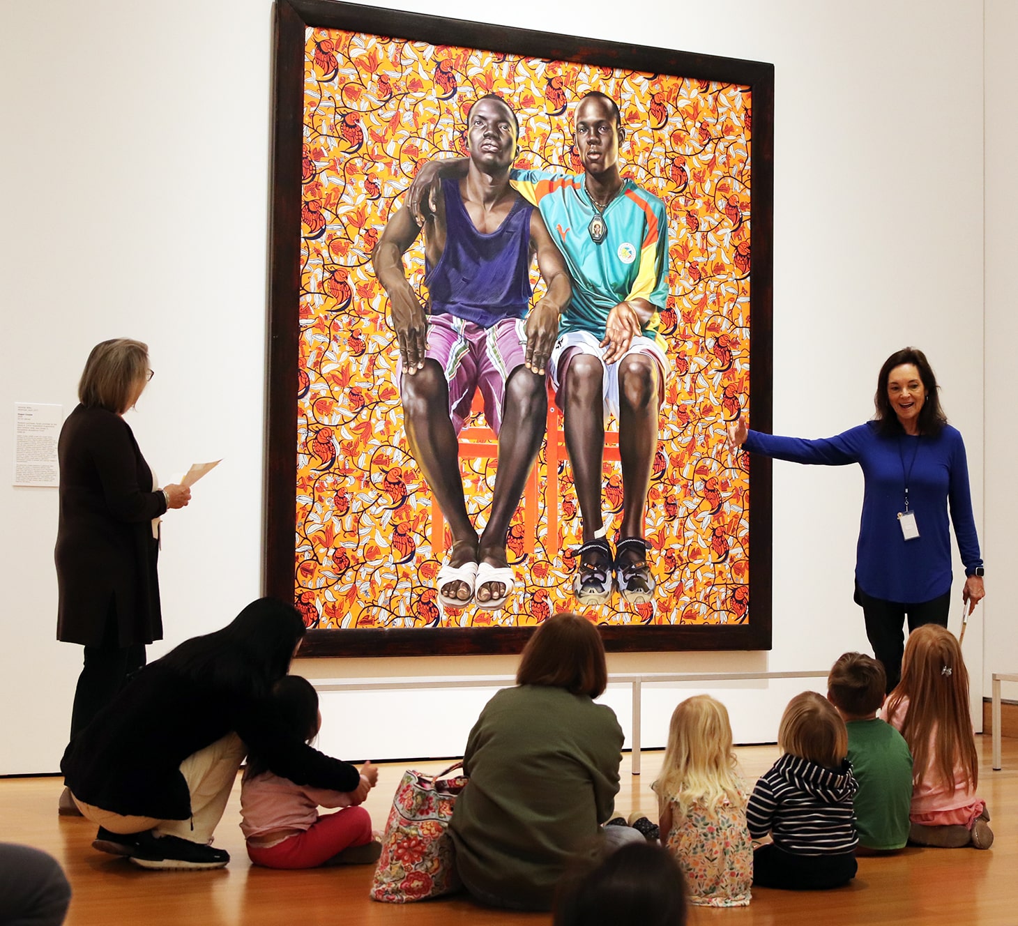 young visitors and their caregivers sitting on the floor in the contemporary gallery listening to a presentation by docents in front of a Kehinde Wiley painting