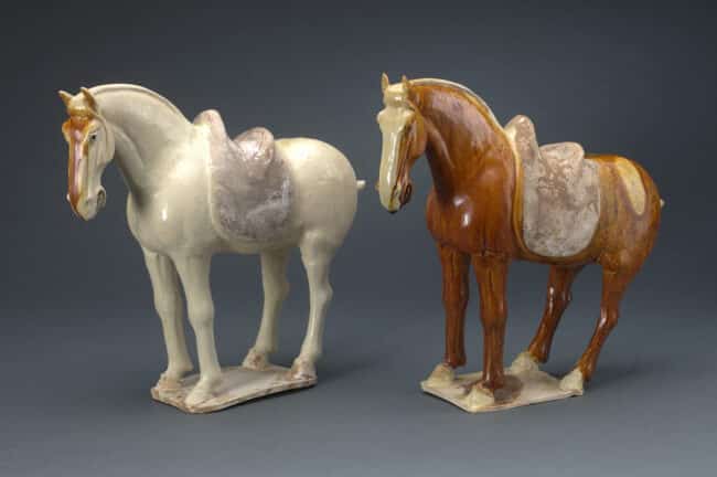 Pair of ceramic horse figurines, one white and one brown, each with detailed saddles and realistic features on a gray background.