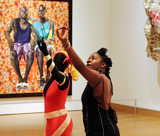 A woman in a black dress stands next to a shadow-like figure in a red dress with gold bracelets. They both raise an arm in a synchronized, expressive gesture. Behind them hangs a large, colorful painting of two men against a vibrant orange, patterned background.