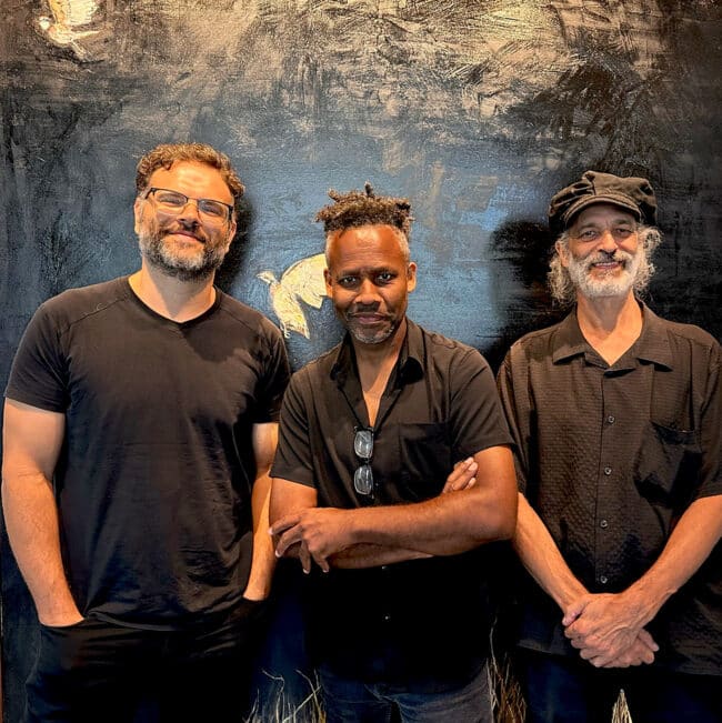 The three members of the band Jazz Life stand side-by-side for a group portrait against a dark, textured painting. All three are casually dressed in black and look directly at the camera casually dressed in black.