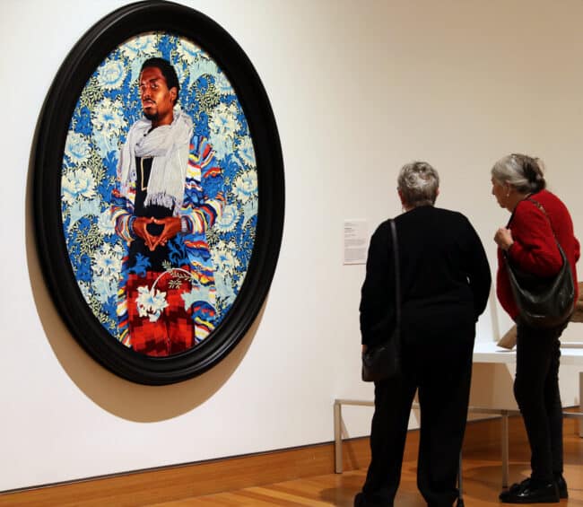 Two women stand in the Harn gallery, looking at a large, oval-framed painting. The vibrant portrait depicts a Black man in a colorful jacket and scarf, positioned against a dense blue and white floral background. The title of the work is "Margaret, Countess of Blessington" by Kehinde Wiley, made in 2018.