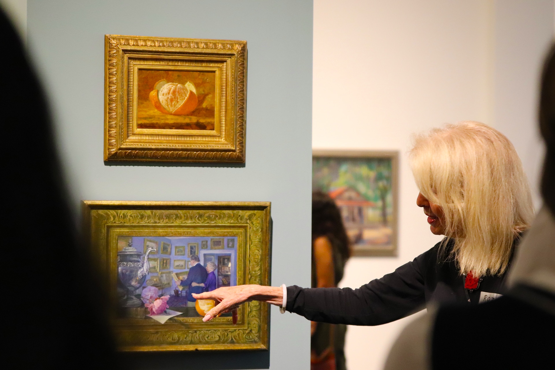 Female docent pointing her index finger towards a painting while looking at it. Her mouth is slightly open as she talks to an audience in the foreground. On the light blue-gray wall to the left, two paintings in ornate gold frames are hung one above the other. The top painting is a small, horizontal still life of a partially peeled orange titled "Orange" by Frank Henry Shapleigh. The bottom painting, which the woman is pointing to, is a slightly larger horizontal scene depicting an interior room where two people are looking at walls covered with many framed pictures titled "Priceless Memories: The Vickers Collection" by Christopher Still.