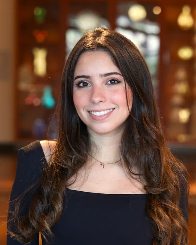 Headshot of Isabela Llovet Lopez, a young woman with long, dark, wavy hair, smiling at the camera. She is wearing a black top with a square neckline and a thin silver necklace. The background is softly blurred shelves of sculptures in the Asian Gallery at the Harn Museum of Art.