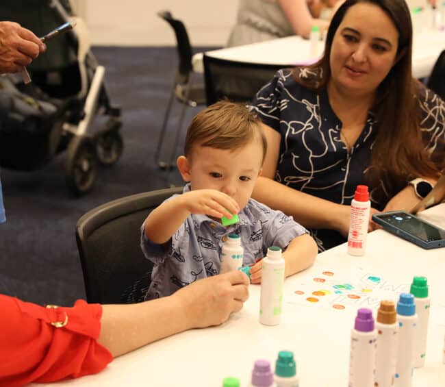 A young boy sits at a white table during an art activity, focused on opening a colorful dot marker. A woman sits beside him, watching with a smile, while various art supplies are spread across the table.