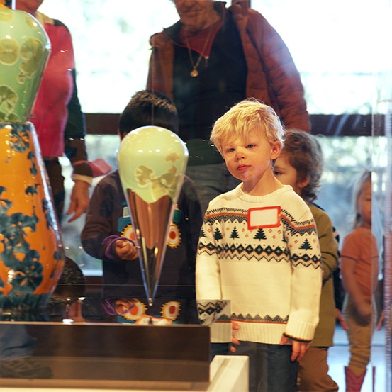 A young blonde child wearing a patterned sweater stands at the Harn Museum of Art, looking closely at a colorful glass sculpture inside a display case. Other children and adults gather in the background, viewing the artwork together during this family-friendly gallery visit.