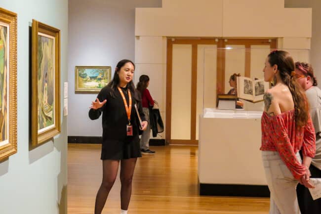 Photograph of a young lady talking while gesturing at a painting in front of a small crowd.